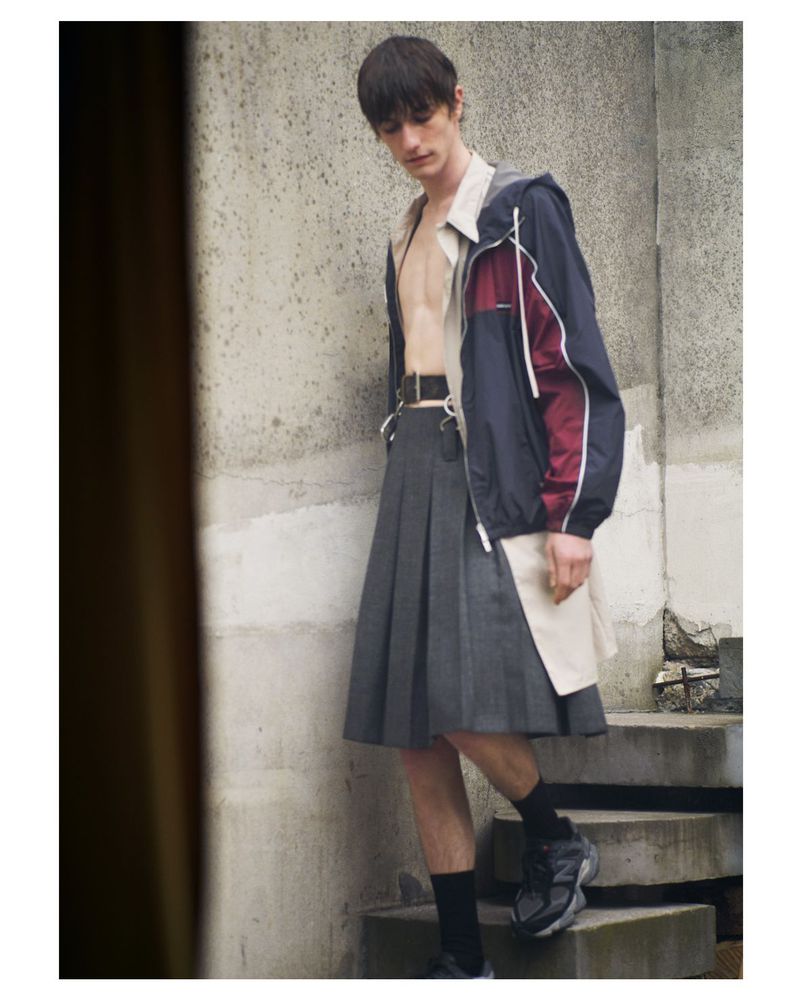 Male fashion model posing outdoors wearing a navy and burgundy hooded jacket with a gray kilt, black socks, and black athletic shoes.