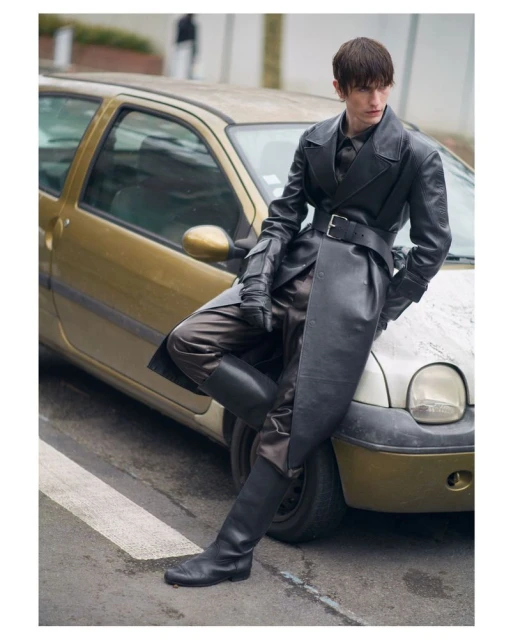 Young adult man in a long black leather coat and boots leans against a gold-colored compact car on a city street with a building background.