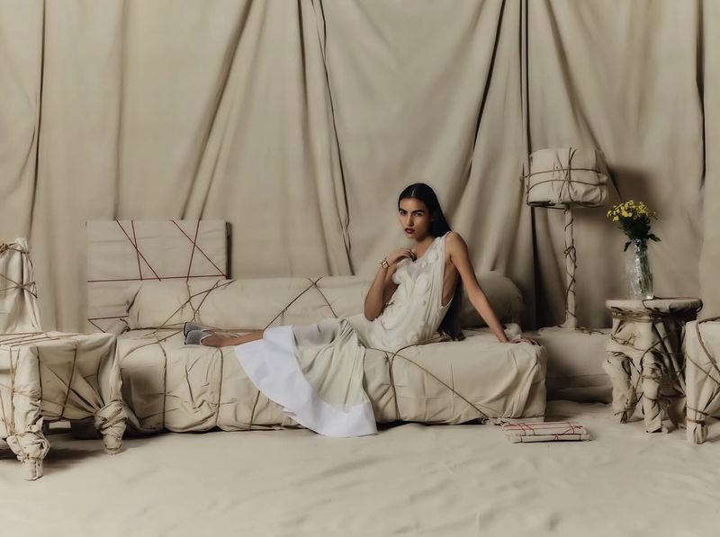 Fashion shot of a young woman in a white dress reclining on furniture wrapped in fabric with thin rope, set against a fabric background.