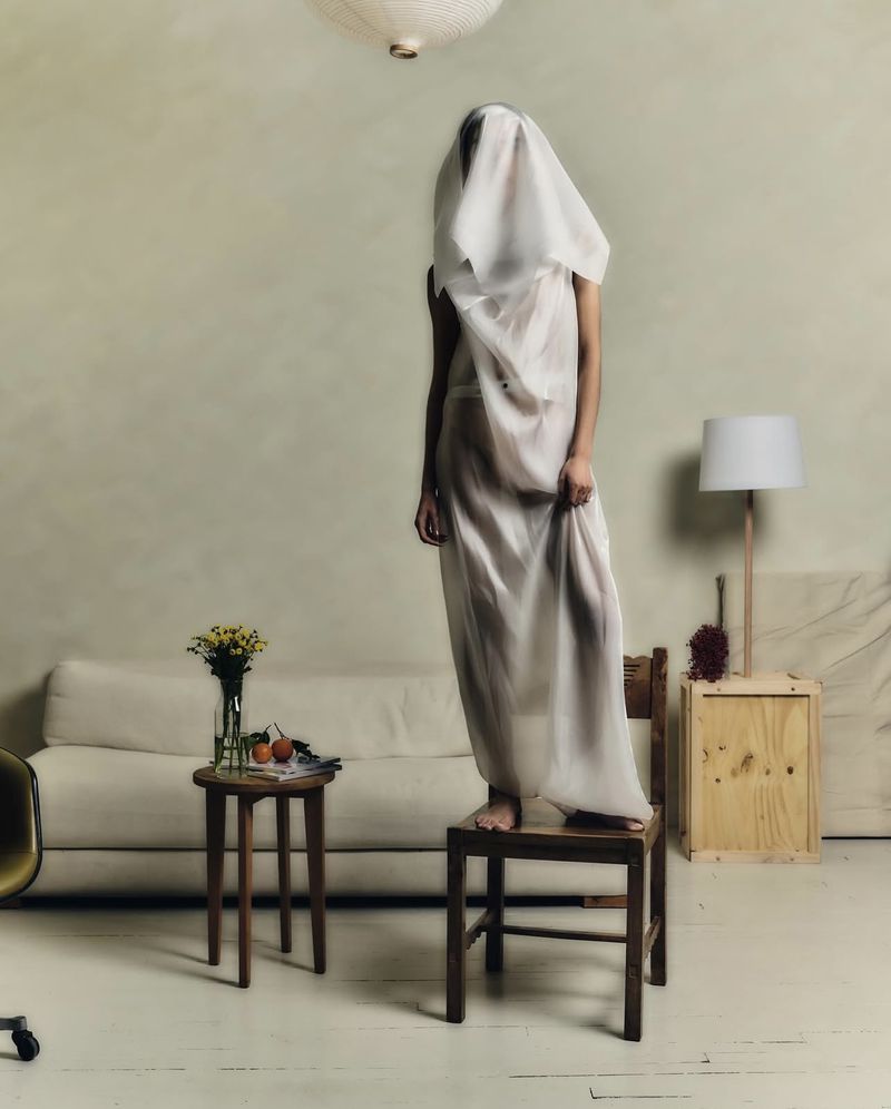 Model standing on a wooden chair draped entirely in translucent white fabric inside a minimal room with a sofa and lamp.