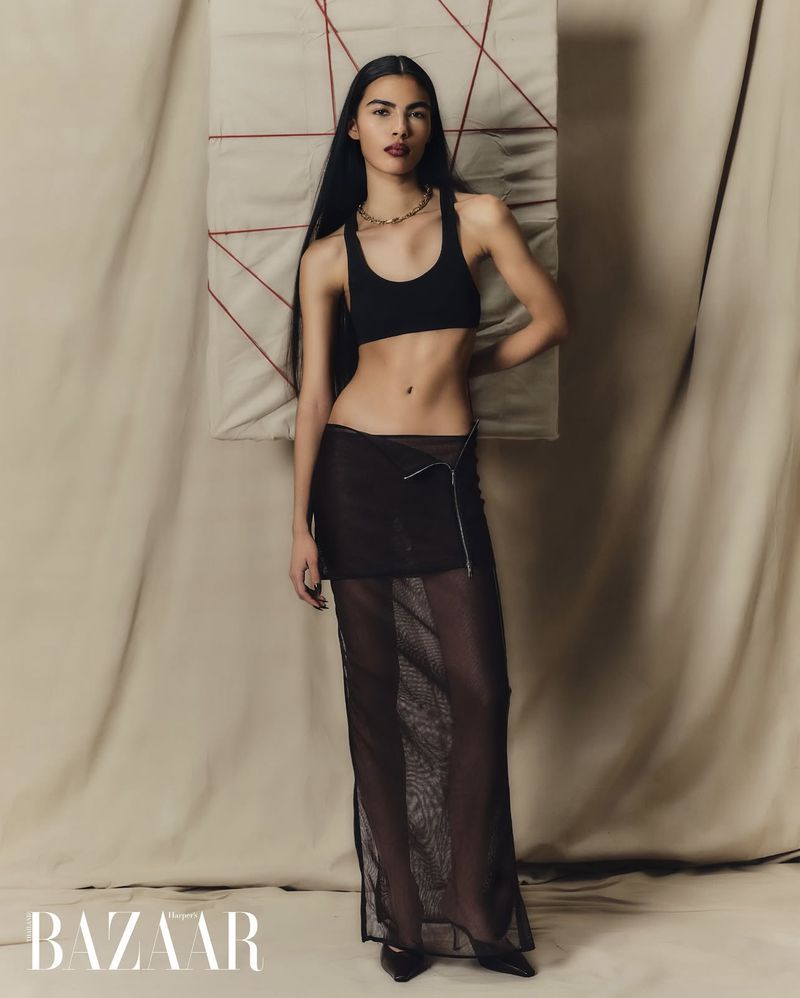 Full-length shot of a fashion model in a black sports bra and mesh skirt posing against a neutral backdrop and artwork.