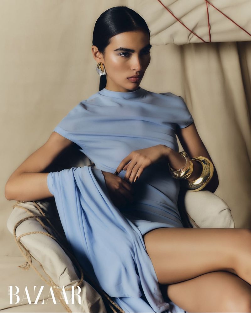 Fashion editorial shot of a woman with dark hair wearing a blue dress and gold cuff bracelet, posing in a draped chair in studio.