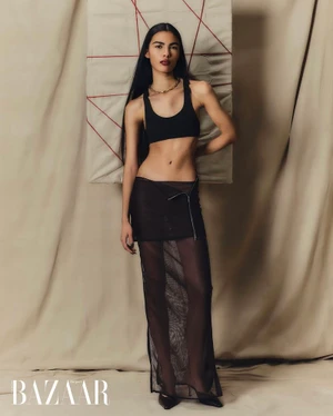 Model with long dark hair wearing a black crop top and sheer maxi skirt standing against a draped fabric backdrop.