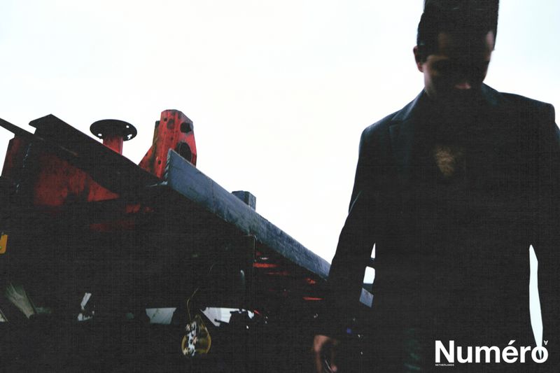 A man in a dark jacket poses near industrial equipment, featuring the 'Numéro' magazine logo, with diffused lighting and a slightly obscured view.