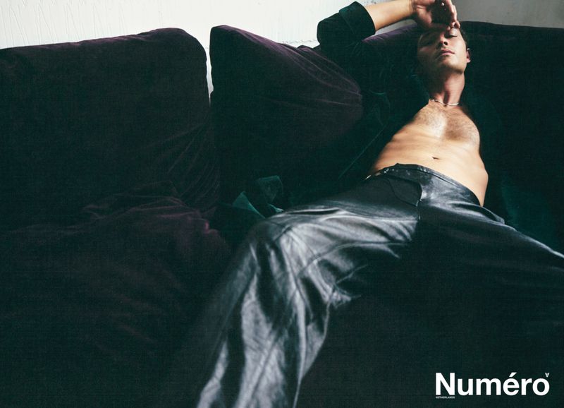 Adult male reclining on a purple velvet couch wearing black pants and a green velvet jacket, indoor setting with neutral lighting.