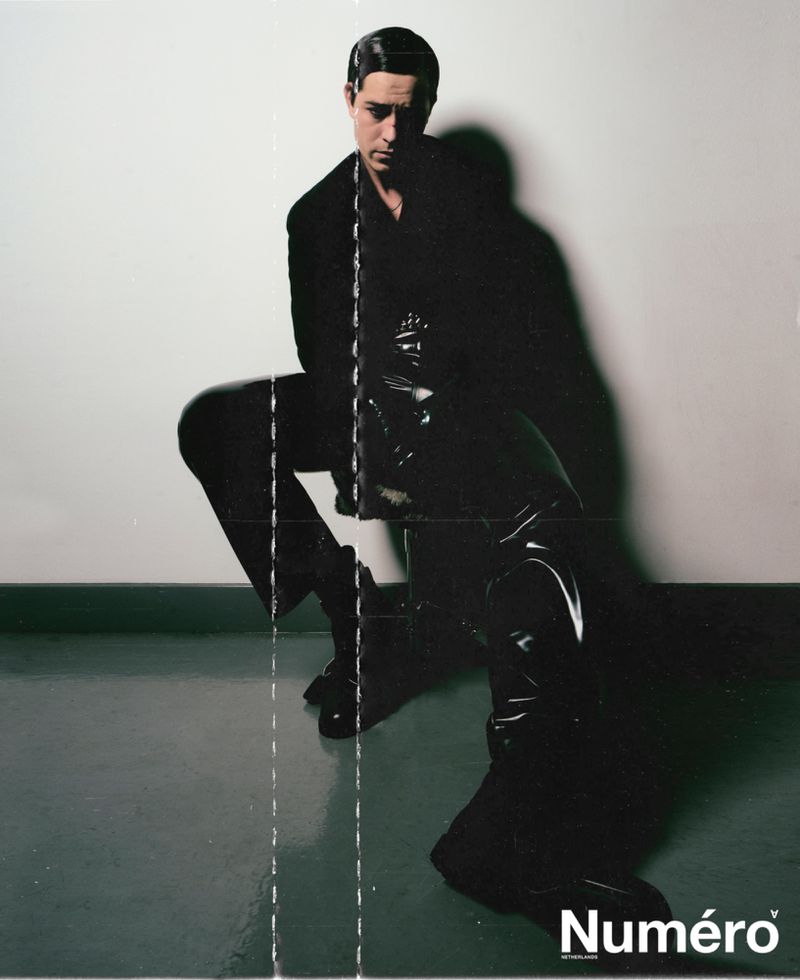 Fashion portrait of a man in a dark suit and shiny boots, posing in front of a plain wall with magazine logo.
