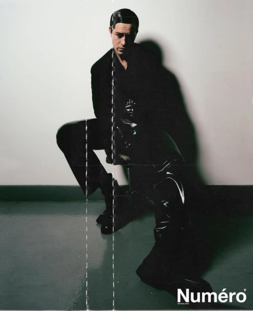 Fashion portrait of a man in a dark suit and shiny boots, posing in front of a plain wall with magazine logo.