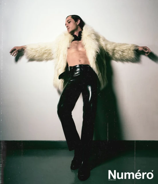 A model strikes a pose, wearing shiny black pants, no shirt, and an open fur coat. The backdrop is a minimalist white wall and floor.