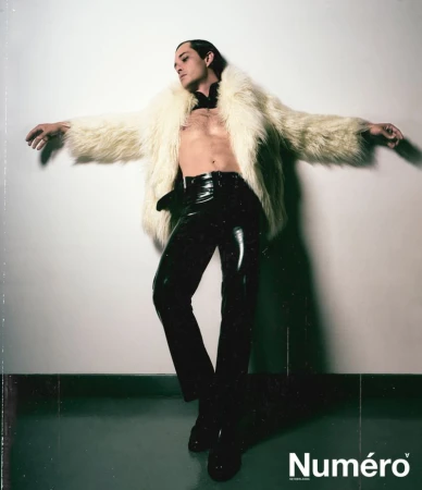 A model strikes a pose, wearing shiny black pants, no shirt, and an open fur coat. The backdrop is a minimalist white wall and floor.