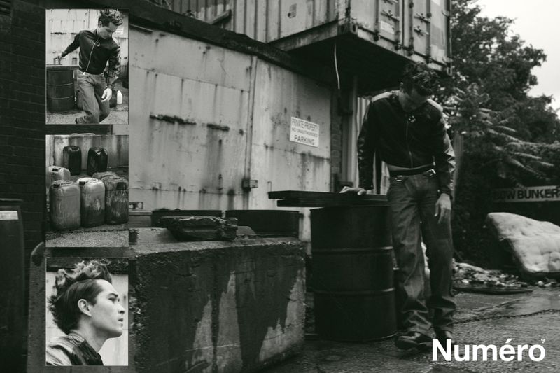 Black and white fashion shot of a man in a leather jacket, standing near barrels, posing next to an industrial shipping container.