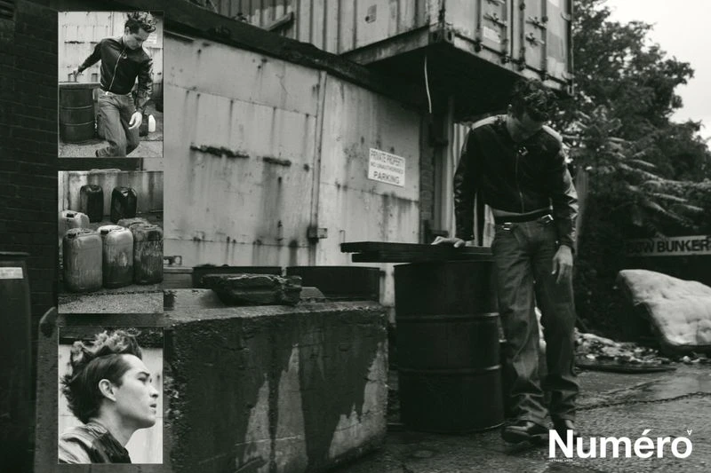 Black and white fashion shot of a man in a leather jacket, standing near barrels, posing next to an industrial shipping container.