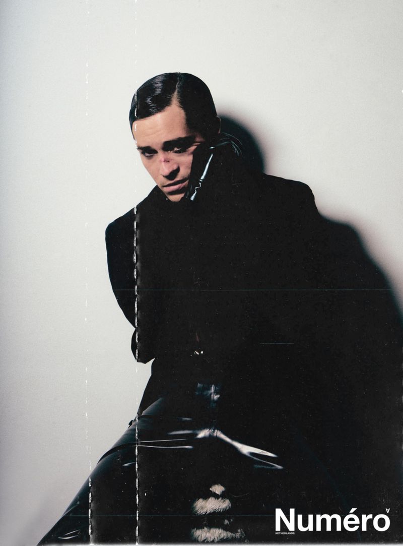 A light-skinned male model with slicked-back hair wearing a black coat and leather pants sits in front of a white background.