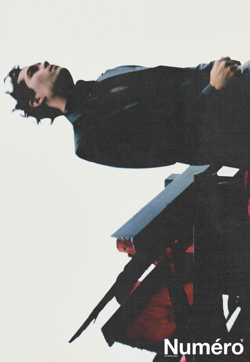 Fashion editorial featuring an adult male model in dark jacket, lying on an abstract structure against a white background.