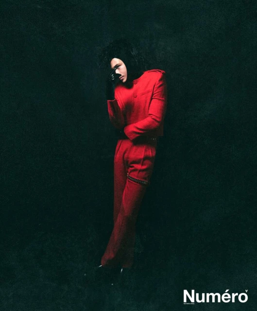Portrait of a person in a red suit and black glove standing in a dim studio. Dark background and "Numero" magazine logo at the bottom.