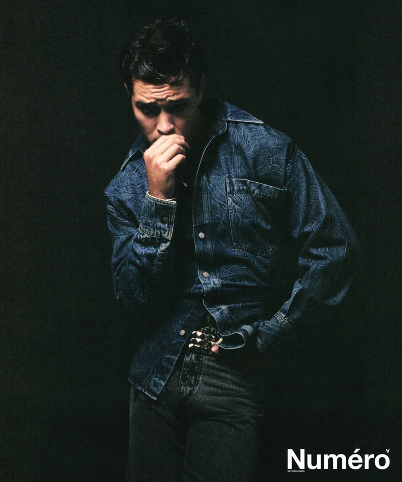 Brooding male fashion model in a blue denim jacket and gray jeans strikes a pensive pose against a stark black background in a photo studio.