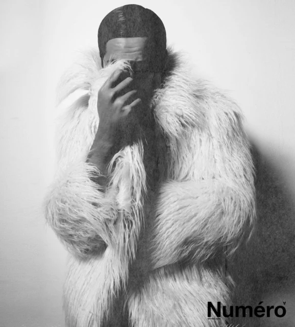 Monochromatic portrait of a person wearing a white faux fur coat, covering their face, and the "Numéro" logo in the corner.