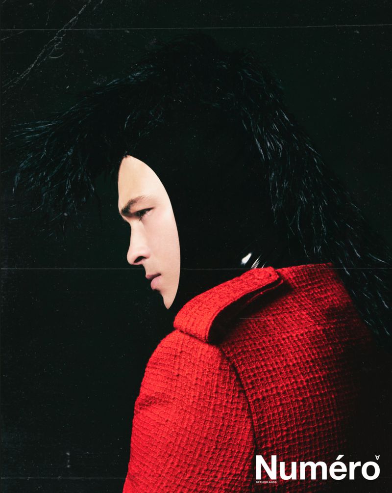 Fashion shot: A model in profile wearing a red jacket and a black hood with feathery details against a dark background for Numero magazine.