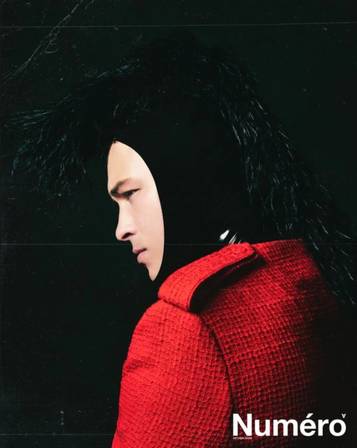 Fashion shot: A model in profile wearing a red jacket and a black hood with feathery details against a dark background for Numero magazine.