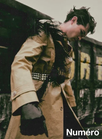 Fashion editorial shot of a young adult male in a tan trench coat, posing against a grungy, urban backdrop with the magazine logo visible.