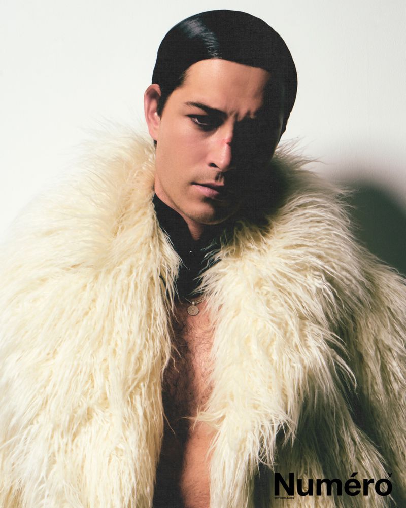 Man in a cream-colored fur coat against a bright white backdrop, looking at the camera, with his chest exposed and styled hair.