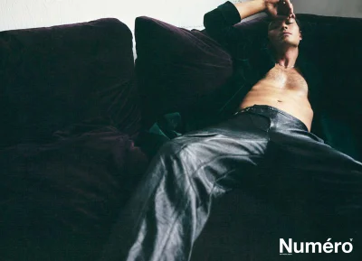 Shirtless male model reclining on a dark couch wearing dark leather trousers for Numero Homme Netherlands by Bruno Unikowsky.