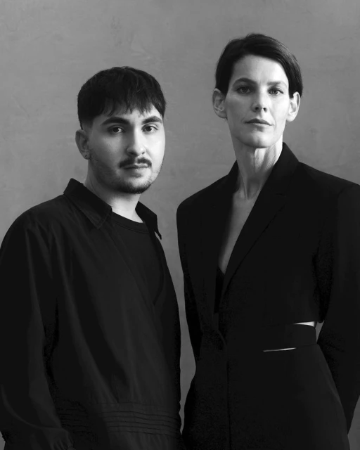 Black and white studio shot of two adults: a man with dark hair and a woman with short hair. Both are in dark clothing against a grey background.