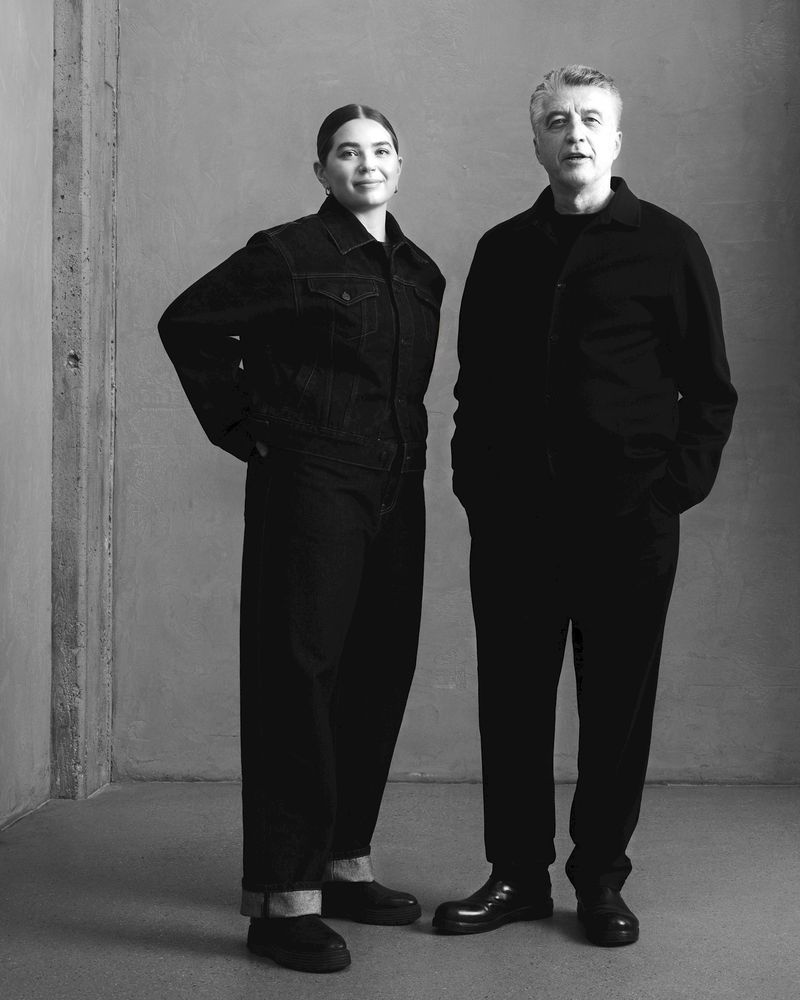 Monochrome portrait of a stylish couple in coordinating black outfits, standing indoors with a neutral grey backdrop and cement wall.
