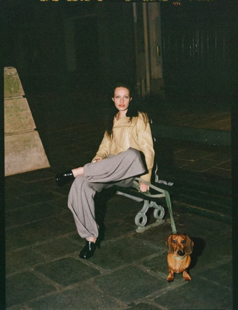 Adult female in a beige jacket and wide-legged pants sits on a bench at night with a brown dachshund dog on a leash.