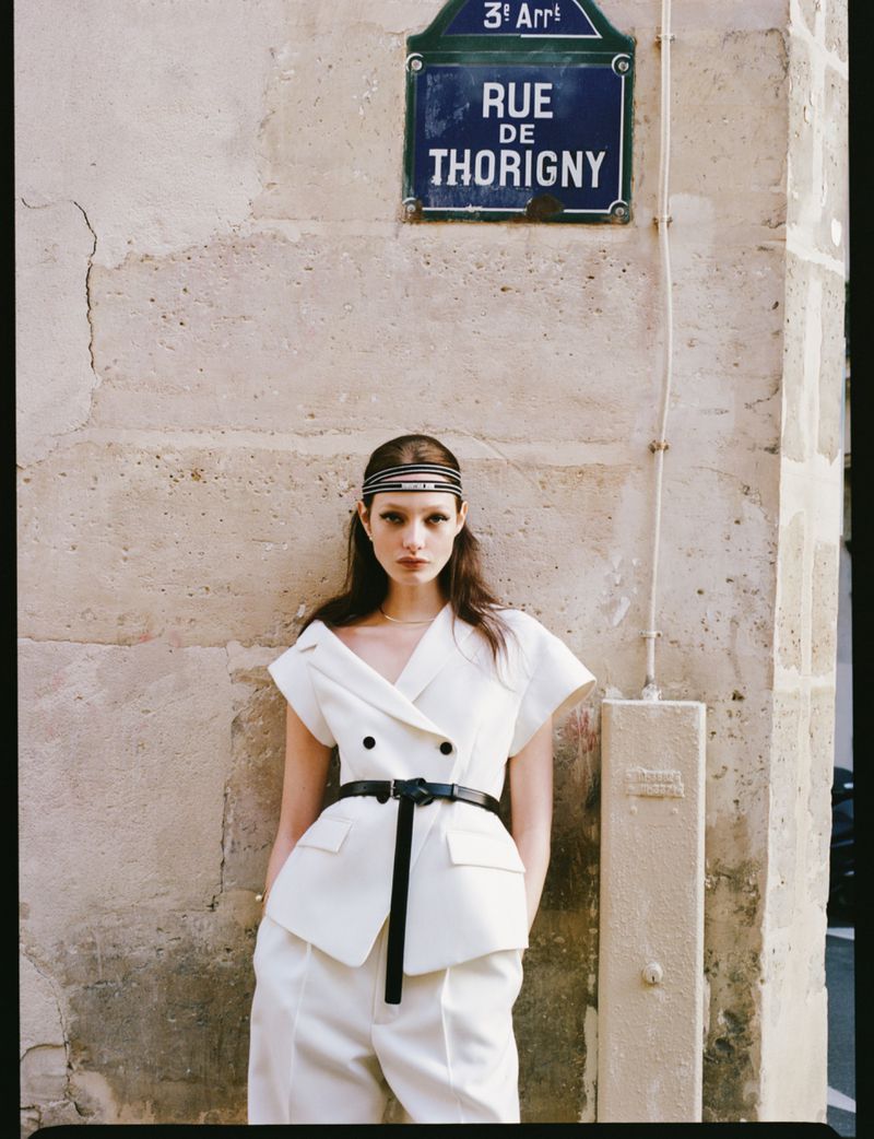 Fashionable woman in a white suit poses against a textured city wall with a street sign, showcasing urban fashion and understated elegance.