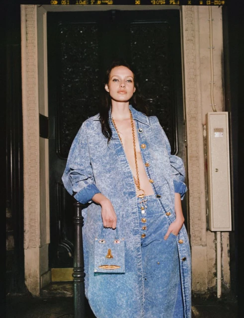 A woman poses in a long denim outfit with a handbag in an urban setting, illuminated by warm lighting against a dark doorway.