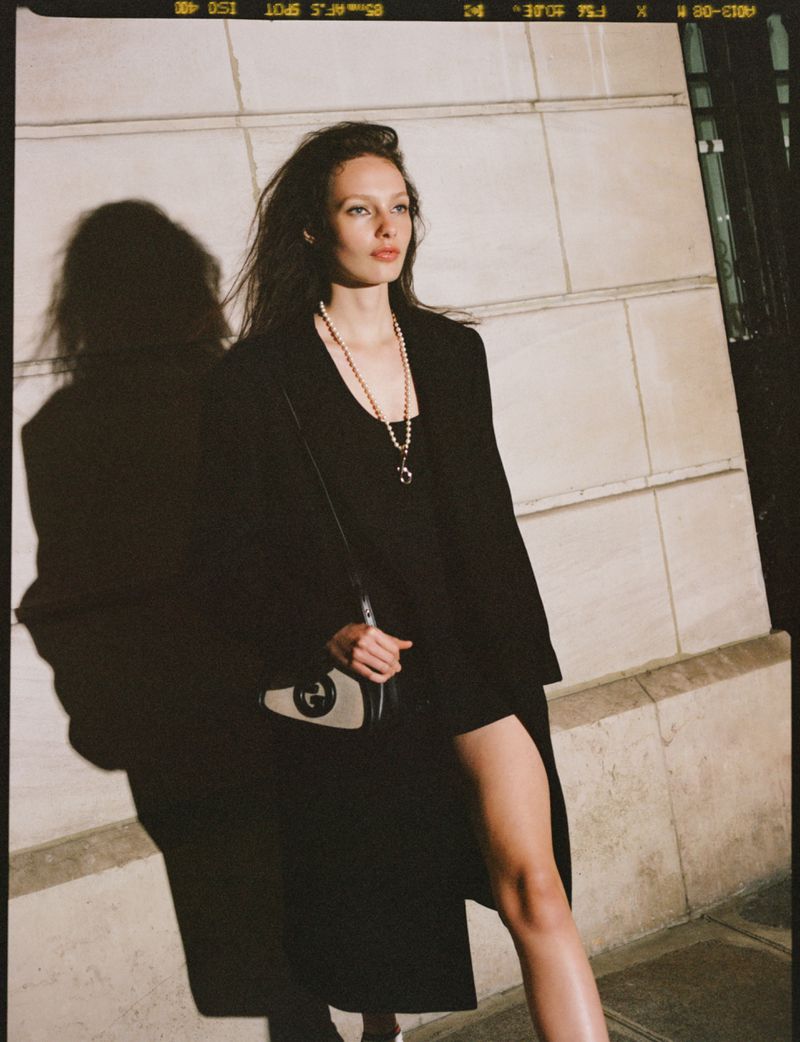 Model in a black coat with a pearl necklace and handbag posing against an exterior stone wall for Vogue Mexico.