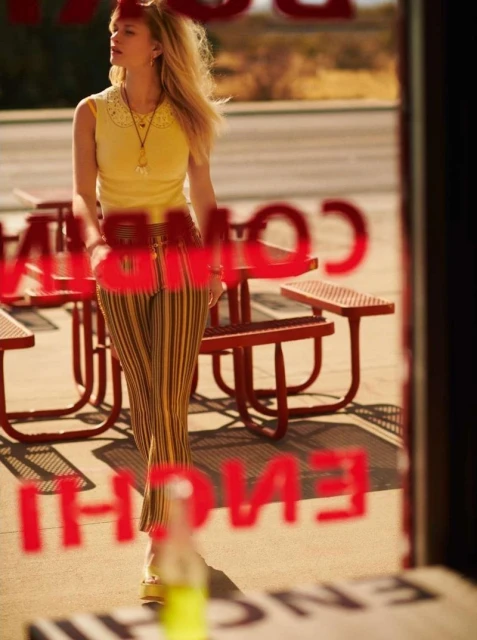 Young woman in yellow top and striped pants stands outside a diner with red picnic tables, viewed through a glass window with red text.