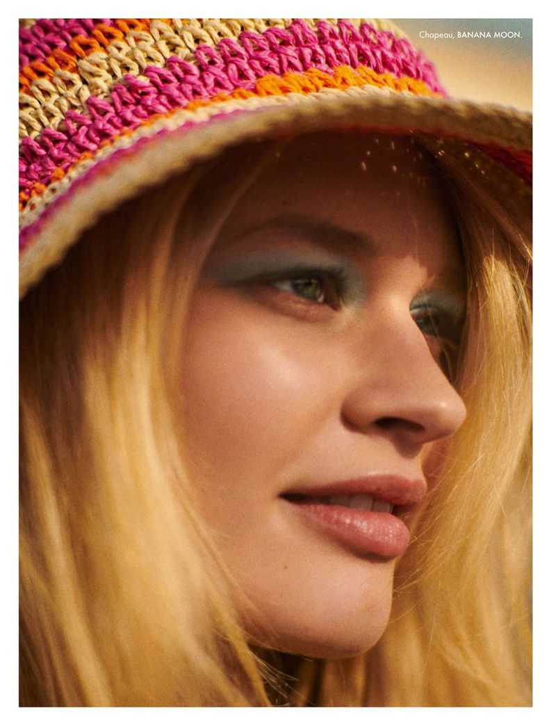 Close-up studio shot of a woman with blond hair wearing a colorful straw hat and blue eye shadow, looking off to the side.