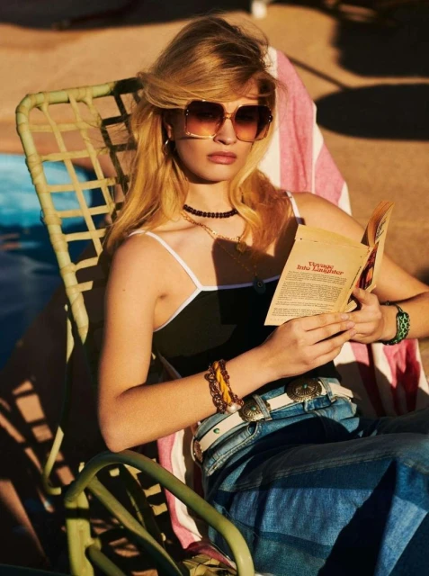 A blonde woman with sunglasses sits in a chair, reading a book by a pool in a bright, sunlit outdoor setting. She wears jewelry and jeans.
