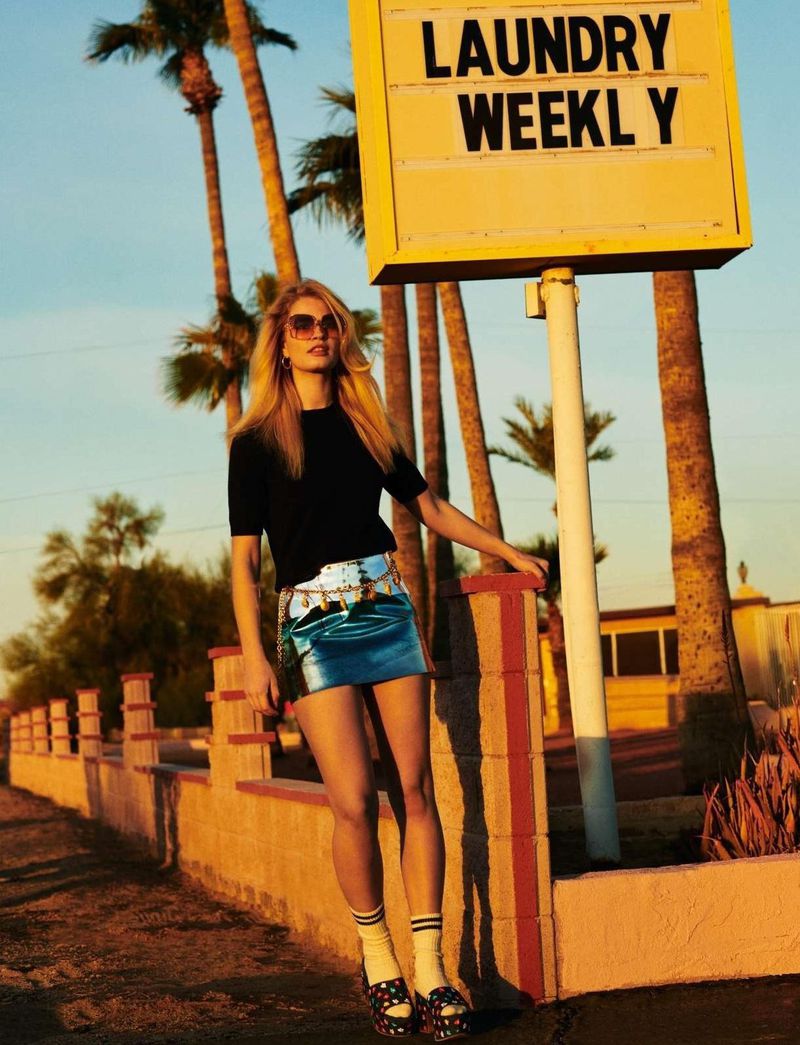 Blonde woman in sunglasses, black top, and blue skirt posing next to a vintage laundry sign and low wall against a backdrop of palm trees.