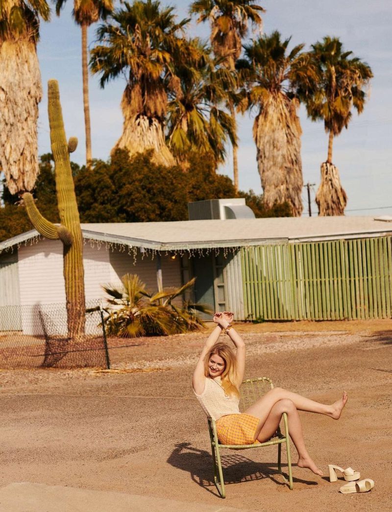 Young woman in yellow gingham shorts sits in a chair outdoors near palm trees and desert plants, enjoying sunny desert weather.