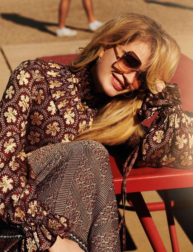 Woman in patterned top and pants sitting in sunlight outdoors. Medium shot featuring neutral earth tone and red colors. Fashion photography.