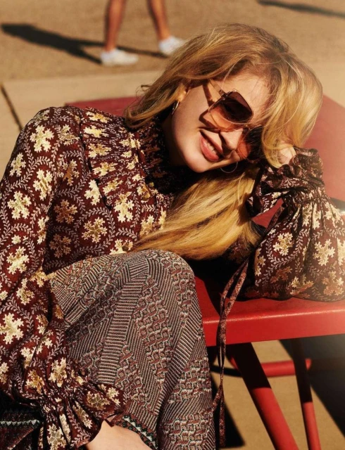 Woman in patterned top and pants sitting in sunlight outdoors. Medium shot featuring neutral earth tone and red colors. Fashion photography.