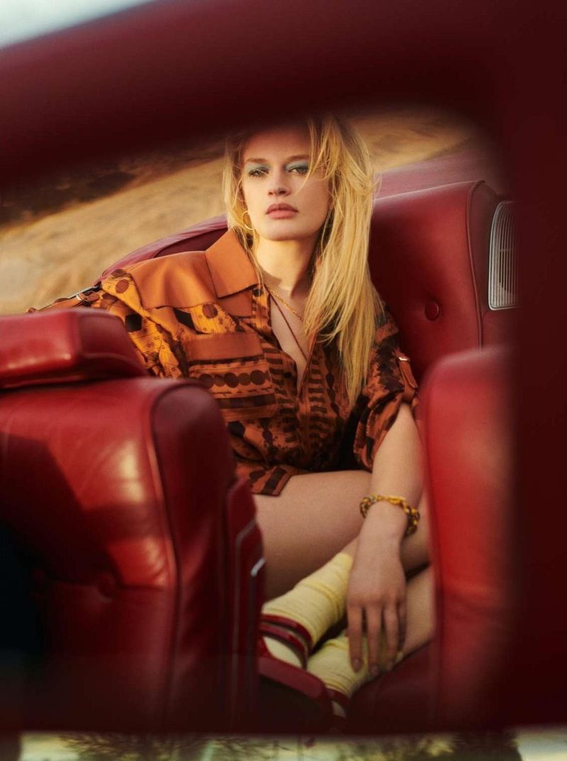 Woman with long blonde hair, wearing an orange patterned shirt, sits in red car seats outdoors in warm light. Fashion shot.