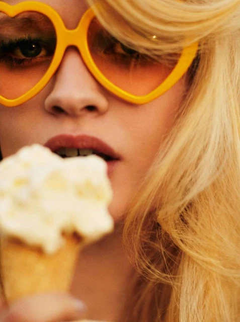 Close-up of an adult female model with blonde hair eating ice cream, wearing heart-shaped yellow sunglasses. Summery and vibrant mood.