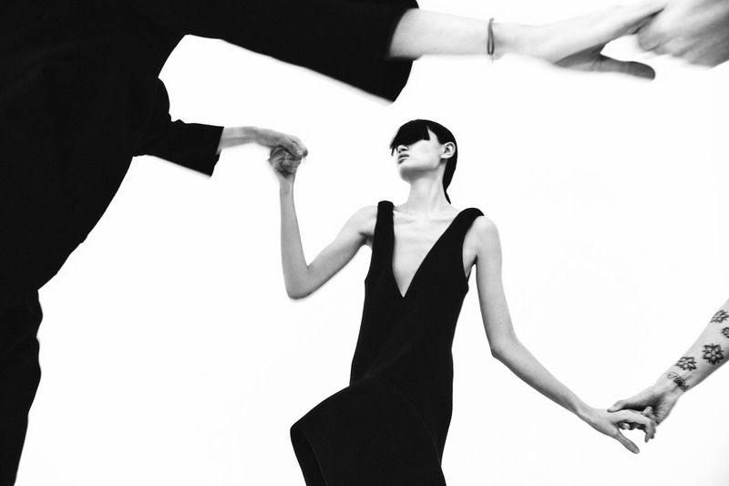 Monochrome fashion shot of a thin model in a black dress, surrounded by hands, against a bright white background, conveying a minimalist aesthetic.