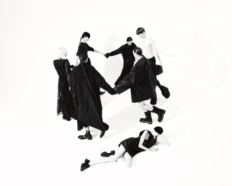 Seven fashion models wearing dark clothing in a circle in a bright studio. One model is lying down. Fashion and studio photography.