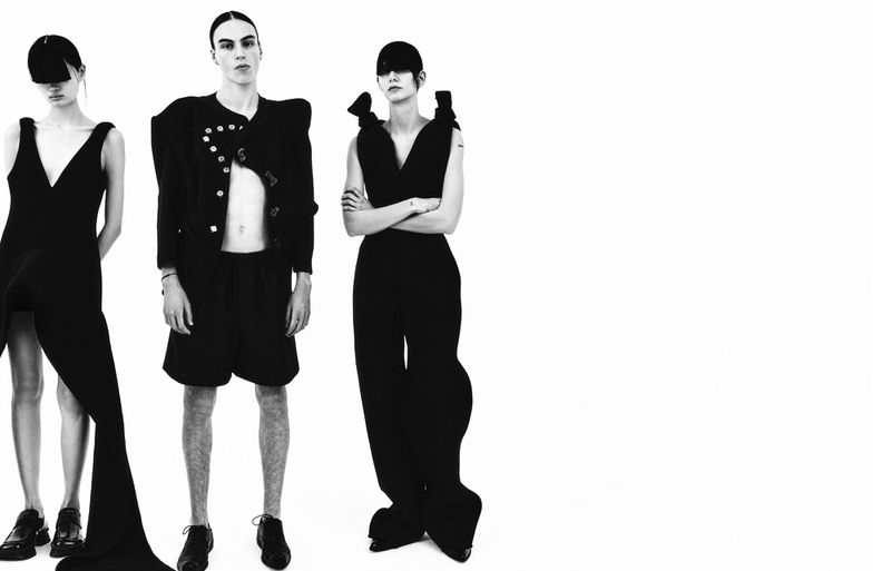 Three fashion models in black clothing pose against a white backdrop, showcasing avant-garde style and minimalist aesthetic in a studio setting.