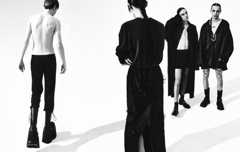 Black and white fashion shot of four slender adults showcasing dramatic dark clothing, combat boots, and edgy poses in a stark studio.