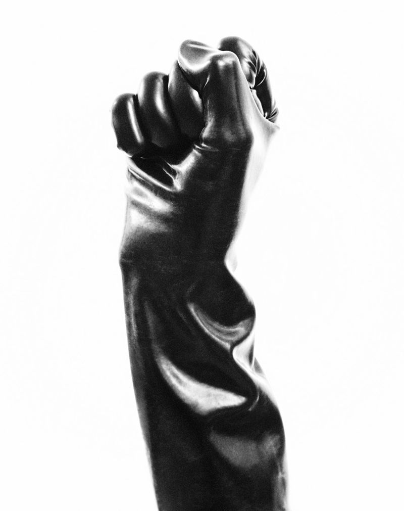 Black latex gloved fist raised in a sign of strength and solidarity against a bright white backdrop in this studio-lit black and white photograph.