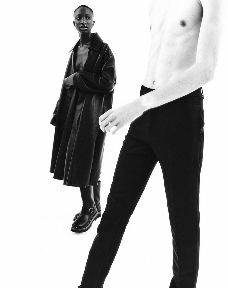 Monochromatic fashion shot features two slender figures against a bright background: one in a long coat and the other shirtless.
