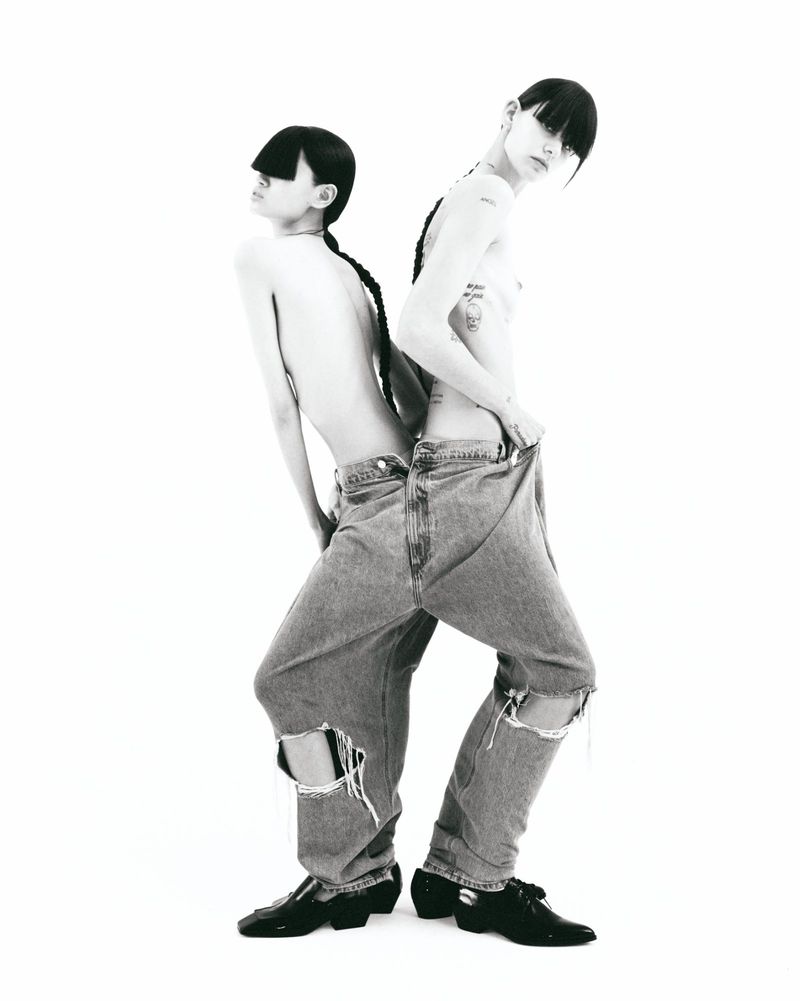 Two shirtless, slender young adults with dark braided hair, posed back-to-back. Both are wearing ripped jeans and patent leather shoes, in studio.