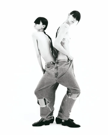 Two shirtless, slender young adults with dark braided hair, posed back-to-back. Both are wearing ripped jeans and patent leather shoes, in studio.