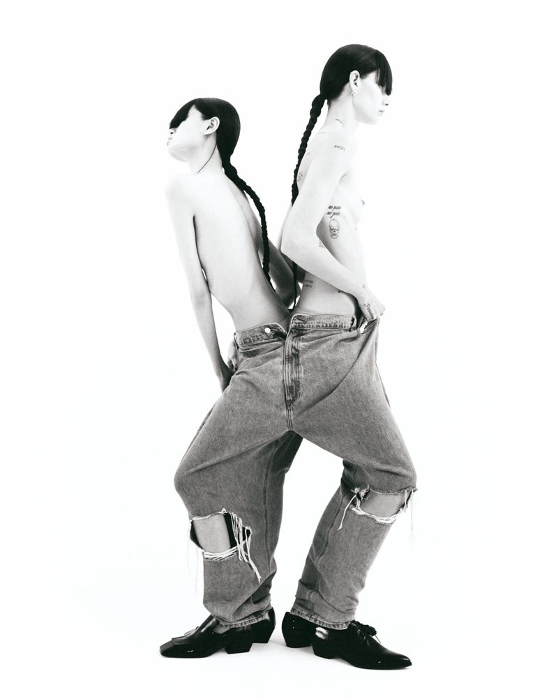 Two shirtless adults in ripped denim stand back-to-back against a white backdrop, showcasing a minimalist fashion concept.