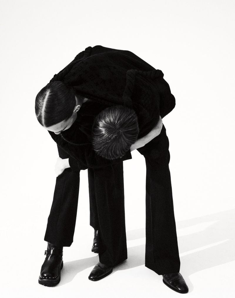Two people in black outfits bend forward against a plain white background in a fashion editorial shot with clean, minimalist composition.